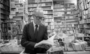 foto em preto e branco do autor folheando um livro entre as bancadas de livros