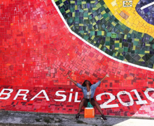 escritora sentada com pernas e braços abertos em frente a uma parede revestida de mosaico de ladrilhos de fundo vermelho onde se lê Brasil 2010 na parte de baixo e uma parte da bandeira brasileira na parte de cima